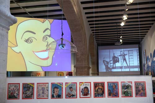 Vista de la exposición que muestra: dos vídeos, una lámpara de diseño de la época y una serie de revistas Time.