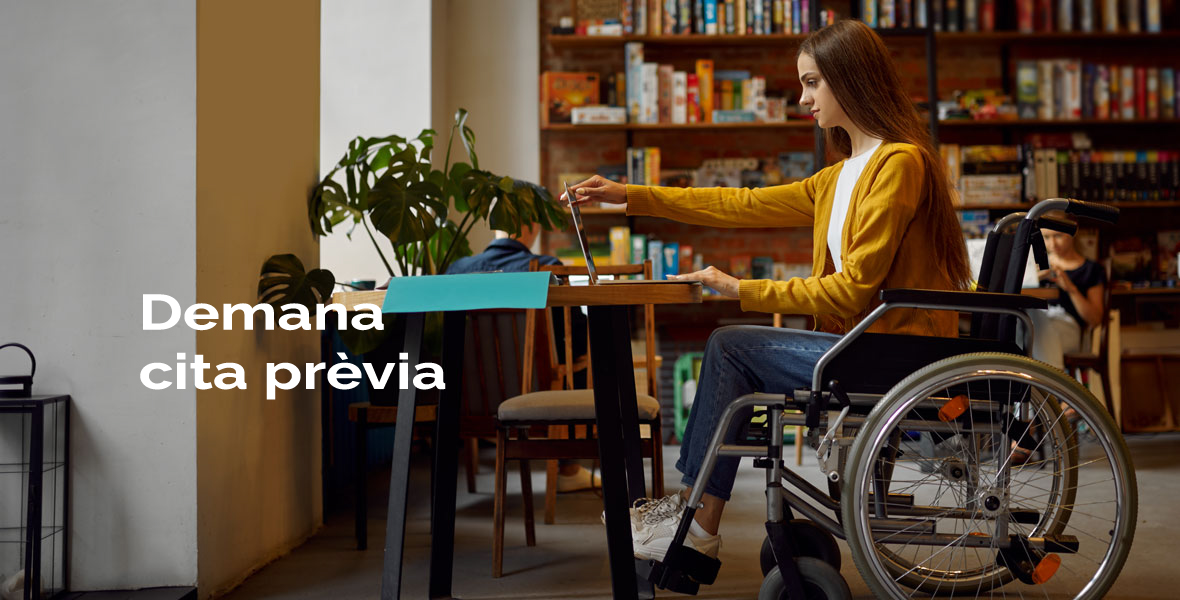 Dona en cadira de rodes mirant un ordinador portàtil en una taula i al fons prestatgeria amb llibres. Hi ha un retole de 
