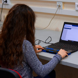 Recursos d'accessibilitat digital - Imatge d'una dona invident asseguda en una cadira i sobre una taula està treballant amb un ordinador adaptat