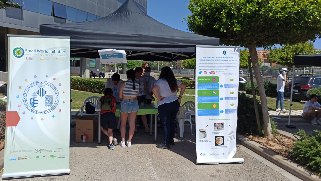 Estand de Micromón a l’aire lliure amb pòsters de Small World Initiative i alumnat consultant materials.