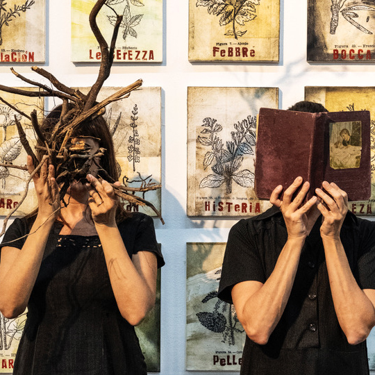 Dos mujeres con la cara tapada. Una  por ramas y la otra por un libro
