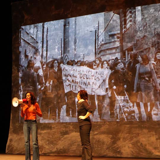Two women on stage and, behind them, a projection of a demonstration