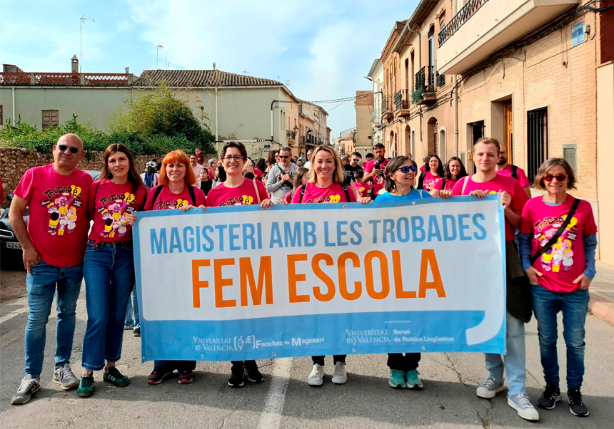 Un grup de docents universitaris, de dones i homes amb la samarreta de les Trobades 2025, sostenen i posen amb una pancarta pel carrer amb el lema 