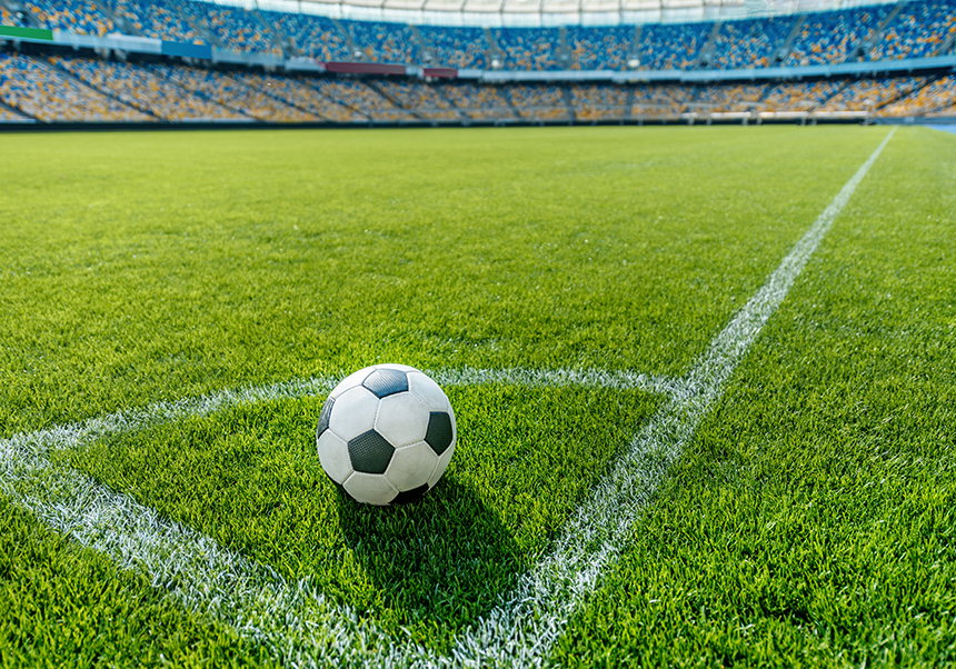 Un baló al córner d'un camp de futbol amb la resta del camp i la grada de fons.