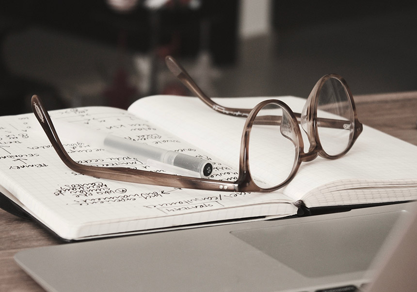 A pair of eyeglasses and a pen can be seen on a notebook with some scientific writing