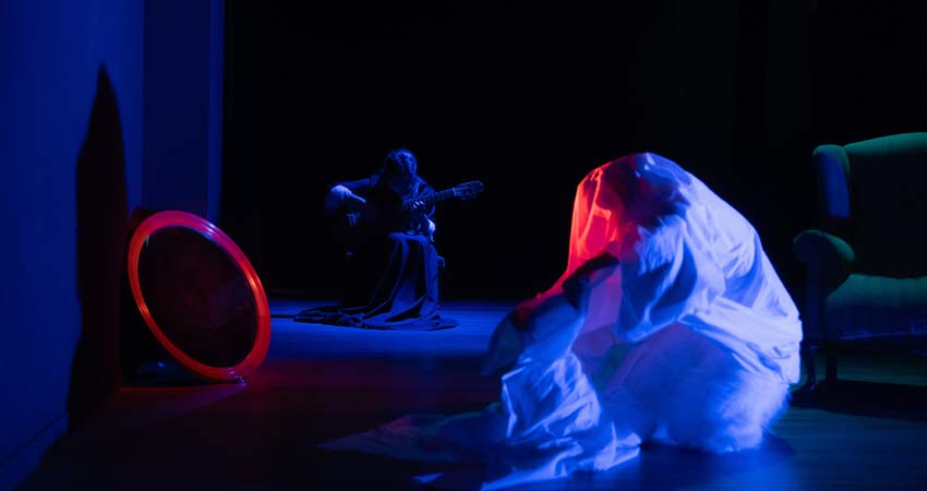 Two people on a stage, one playing the guitar, the other wrapped in a sheet