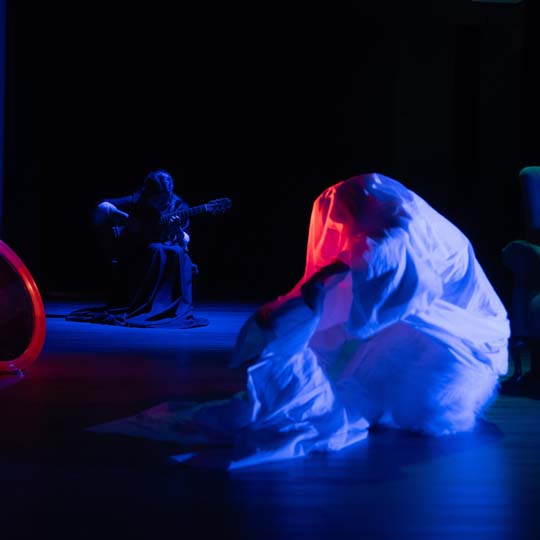 Two people on a stage, one playing the guitar, the other wrapped in a sheet