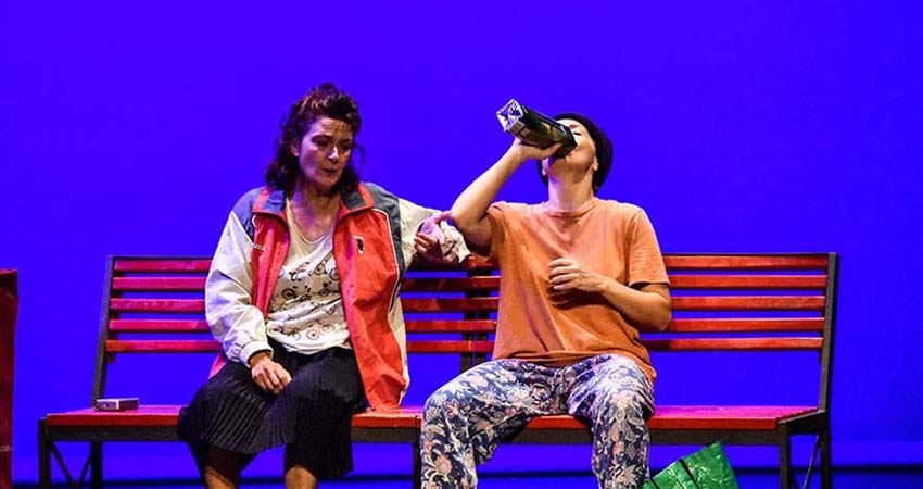 Two actresses sitting on a bench one of them drinking