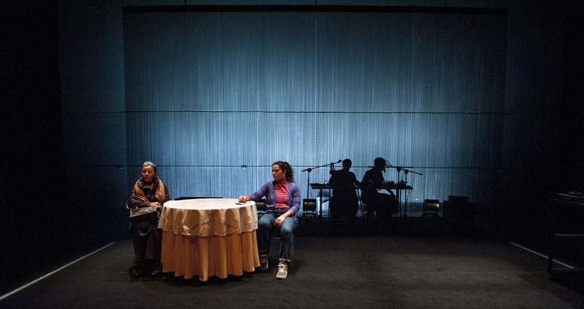 Two actresses in front of a table and two musicians in the background