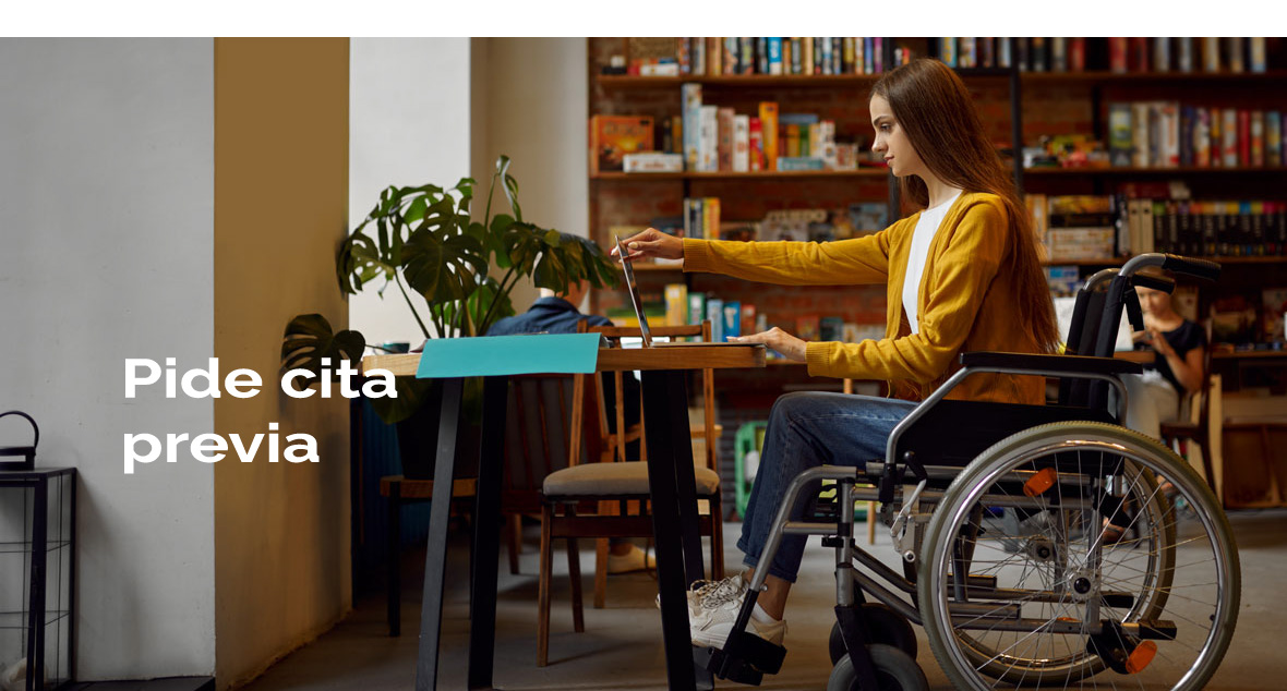 Mujer en silla de ruedas mirando un ordenador portátil en una mesa y al fondo estantería con libros. Hay un rotulo de 