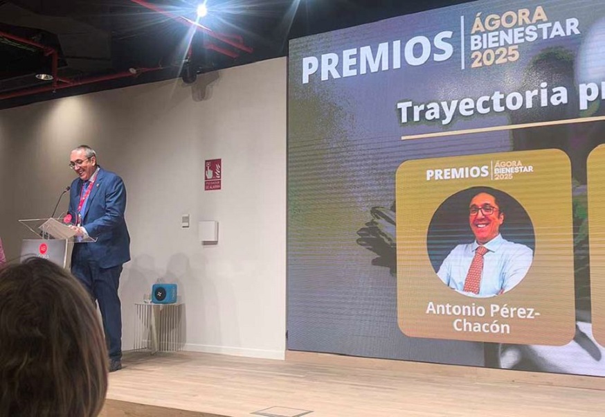 A speaker at a lectern during the Ágora Bienestar 2025 Awards, with a screen showing the professional career award for Antonio Pérez-Chacón.