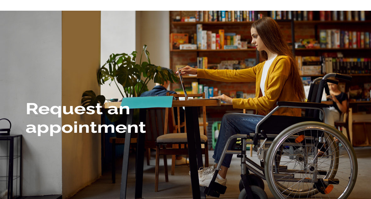 A woman in a wheelchair looking at a laptop on a table with a bookshelf full of books in the background. There is a sign 